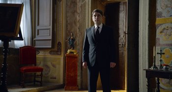 Movie still from “The Two Popes” (2019), directed by Fernando Meirelles – A man in a suit standing in front of a statue; Medium shot, Low angle