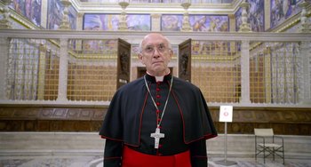 Movie still from “The Two Popes” (2019), directed by Fernando Meirelles – An older man in a red and black outfit with a crucifix in front of him; Close Up shot, Over the shoulder angle