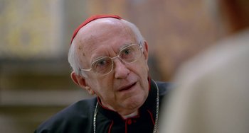 Movie still from “The Two Popes” (2019), directed by Fernando Meirelles – An older man wearing glasses and wearing a red hat; Close Up shot, Over the shoulder angle