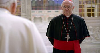 Movie still from “The Two Popes” (2019), directed by Fernando Meirelles – An older man wearing a red and black outfit; Close Up shot, Over the shoulder angle