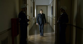 Movie still from “The Two Popes” (2019), directed by Fernando Meirelles – A man in a black suit walking down a hall way; Medium shot, Over the shoulder angle