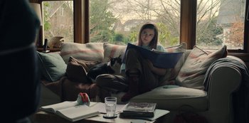 Movie still from “The Unforgivable” (2021), directed by Nora Fingscheidt – A woman sitting on a couch reading a book with a cat sitting on the couch; Medium shot, Over the shoulder angle