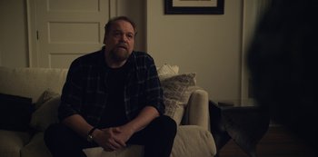 Movie still from “The Unforgivable” (2021), directed by Nora Fingscheidt – A man sitting on top of a couch in a living room; Medium shot, Over the shoulder angle