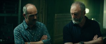 Movie still from “The Vault” (2021), directed by Jaume Balagueró – Two men are sitting in a room and one of them has his arms crossed and one of them has his arms crossed; Close Up shot, Low angle