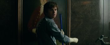 Movie still from “The Vault” (2021), directed by Jaume Balagueró – A man holding a baseball bat in front of a painting; Medium shot, Over the shoulder angle