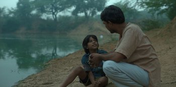 Movie still from “The White Tiger” (2021), directed by Ramin Bahrani – A man and a boy sitting next to a body of water; Medium shot, High angle