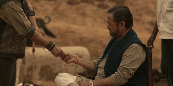 Movie still from “The White Tiger” (2021), directed by Ramin Bahrani – An older man sitting on the ground holding a bracelet; Medium shot, High angle