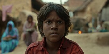 Movie still from “The White Tiger” (2021), directed by Ramin Bahrani – A young boy in a red shirt is looking at the camera; Close Up shot, Low angle