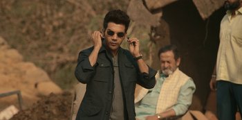 Movie still from “The White Tiger” (2021), directed by Ramin Bahrani – A man in sunglasses and a jacket holding a pair of sunglasses; Medium shot, Low angle