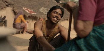 Movie still from “The White Tiger” (2021), directed by Ramin Bahrani – A man sitting on the ground smiling at the camera; Close Up shot, Over the shoulder angle