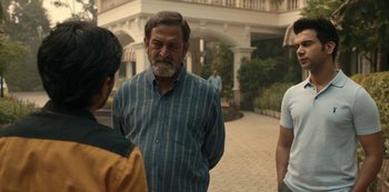 Movie still from “The White Tiger” (2021), directed by Ramin Bahrani – A man standing next to two other men in front of a building; Medium shot, Over the shoulder angle