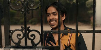 Movie still from “The White Tiger” (2021), directed by Ramin Bahrani – A man standing behind a metal gate with his mouth open; Close Up shot, Over the shoulder angle