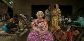 Movie still from “The White Tiger” (2021), directed by Ramin Bahrani – An old woman sitting on a chair in front of a group of people; Medium shot, High angle