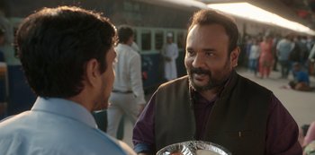 Movie still from “The White Tiger” (2021), directed by Ramin Bahrani – A man talking to another man while holding a plate of food; Medium shot, Over the shoulder angle