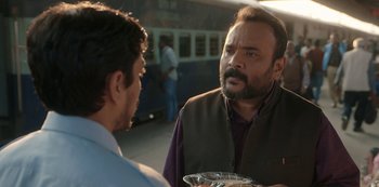 Movie still from “The White Tiger” (2021), directed by Ramin Bahrani – A man holding a plate talking to another man in front of a train; Medium shot, Over the shoulder angle