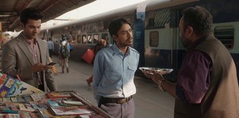 Movie still from “The White Tiger” (2021), directed by Ramin Bahrani – A man holding a plate of food in front of a train; Medium shot, Over the shoulder angle