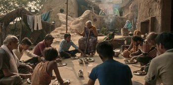 Movie still from “The White Tiger” (2021), directed by Ramin Bahrani – A group of people sitting around a table with food; Wide shot, Over the shoulder angle