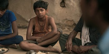 Movie still from “The White Tiger” (2021), directed by Ramin Bahrani – A young boy sitting on the ground next to an older man; Close Up shot, High angle