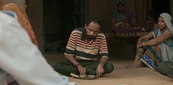 Movie still from “The White Tiger” (2021), directed by Ramin Bahrani – A man sitting on the ground holding a piece of wood; Medium shot, Low angle