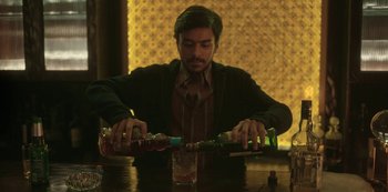 Movie still from “The White Tiger” (2021), directed by Ramin Bahrani – A man sitting at a table with two bottles of alcohol; Medium shot, Low angle