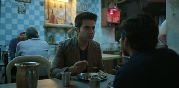 Movie still from “The White Tiger” (2021), directed by Ramin Bahrani – A man sitting at a table with a plate of food in front of him; Medium shot, Over the shoulder angle