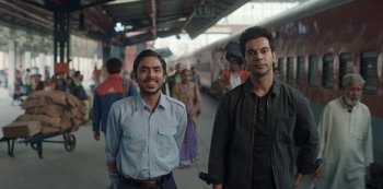 Movie still from “The White Tiger” (2021), directed by Ramin Bahrani – Two men standing next to each other on a train platform; Medium shot, Low angle