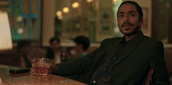 Movie still from “The White Tiger” (2021), directed by Ramin Bahrani – A man sitting at a table holding a glass of wine; Medium shot, Low angle
