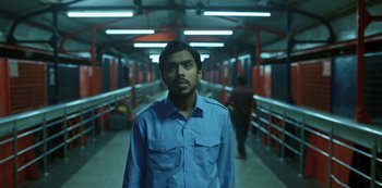 Movie still from “The White Tiger” (2021), directed by Ramin Bahrani – A man in a blue shirt standing in a hallway; Medium shot, Low angle