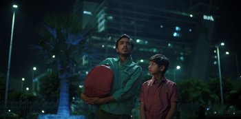 Movie still from “The White Tiger” (2021), directed by Ramin Bahrani – A man and a boy standing next to each other in front of a building; Medium shot, Low angle