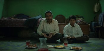 Movie still from “The White Tiger” (2021), directed by Ramin Bahrani – Two men sitting at a table with plates of food on it; Medium shot, Low angle