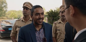 Movie still from “The White Tiger” (2021), directed by Ramin Bahrani – A man in a blue suit and sunglasses standing in front of two police officers; Medium shot, Over the shoulder angle