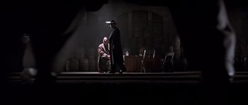 Movie still from “Road to Perdition” (2002), directed by Sam Mendes – A man in a hat is walking on a stage with another man; Wide shot, Over the shoulder angle