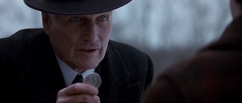 Movie still from “Road to Perdition” (2002), directed by Sam Mendes – An older man wearing a black hat holding a coin in his hand; Close Up shot, Over the shoulder angle