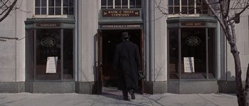 Movie still from “Road to Perdition” (2002), directed by Sam Mendes – A man in a long black coat and a black hat walking out of a bank and trust company; Wide shot, Low angle