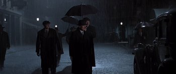 Movie still from “Road to Perdition” (2002), directed by Sam Mendes – A man in a black coat and hat holding an umbrella; Wide shot, Low angle
