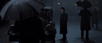 Movie still from “Road to Perdition” (2002), directed by Sam Mendes – A man standing next to an old car in the rain; Wide shot, Over the shoulder angle