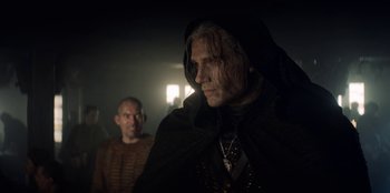 Movie still from “The Witcher” (2019), created by Lauren Schmidt Hissrich – A man in a hooded cloak standing next to another man in a dark room; Close Up shot, Over the shoulder angle