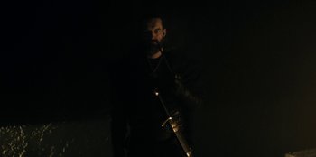 Movie still from “The Witcher” (2019), created by Lauren Schmidt Hissrich – A man with a beard holding a sword in the dark; Medium shot, Low angle