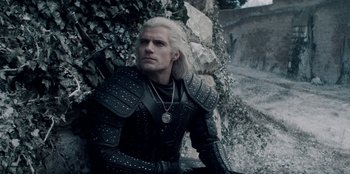 Movie still from “The Witcher” (2019), created by Lauren Schmidt Hissrich – A man in a black leather jacket sitting in front of a rock wall; Medium shot, Over the shoulder angle