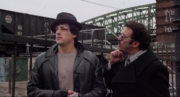 Movie still from “Rocky” (1976), directed by John G. Avildsen – A man in a hat and a man in a leather jacket; Medium shot, Low angle