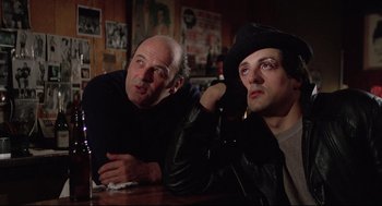 Movie still from “Rocky” (1976), directed by John G. Avildsen – Two men sitting at a table in front of each other; Medium shot, Low angle