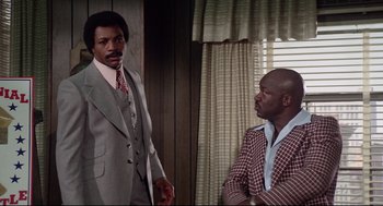 Movie still from “Rocky” (1976), directed by John G. Avildsen – Two men in suits standing next to each other in front of a window; Medium shot, Low angle