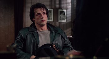 Movie still from “Rocky” (1976), directed by John G. Avildsen – A man sitting in a chair wearing a leather jacket; Medium shot, Over the shoulder angle