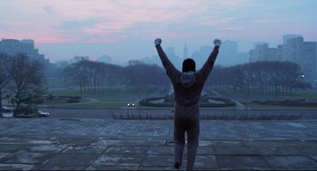 Movie still from “Rocky” (1976), directed by John G. Avildsen – A man standing on a sidewalk with his arms in the air; Wide shot, Low angle