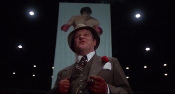 Movie still from “Rocky” (1976), directed by John G. Avildsen – A man in a suit and hat standing in front of a poster; Medium shot, Low angle