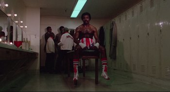 Movie still from “Rocky” (1976), directed by John G. Avildsen – A man sitting on top of a wooden chair in a room; Medium shot, Low angle