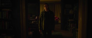 Movie still from “The Woman in the Window” (2021), directed by Joe Wright – A man standing in the dark in front of a living room; Medium shot, Over the shoulder angle
