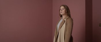 Movie still from “The Woman in the Window” (2021), directed by Joe Wright – A woman wearing a brown coat standing next to a pink wall; Medium shot, Low angle