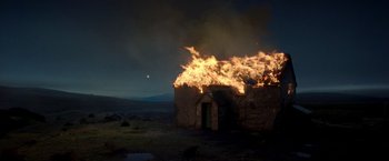 Movie still from “The Wonder” (2022), directed by Sebastián Lelio – An old stone building is burning in the dark; Extreme Wide shot, Low angle
