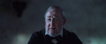 Movie still from “The Wonder” (2022), directed by Sebastián Lelio – An old man with a beard wearing glasses and a bow tie; Close Up shot, Over the shoulder angle